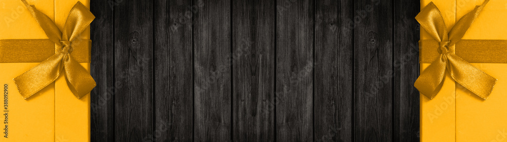Fototapeta premium Festive christmas / celebration / birthday background banner panorama - Yellow gift boxes with golden ribbon, isolated on dark black rustic wooden boards wall / wood texture, with space for text