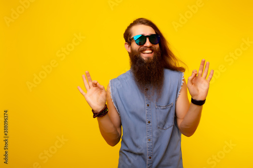 Young smiling man with long hair and beard wearing casual denim t-shirt and sunglasses doing ok gesture with smiling face. He standing over yellow background with copy space.