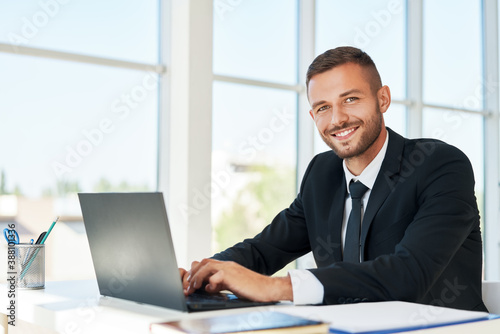 Handsome smiling businessman in elegant suit working on laptop in a bright modern office