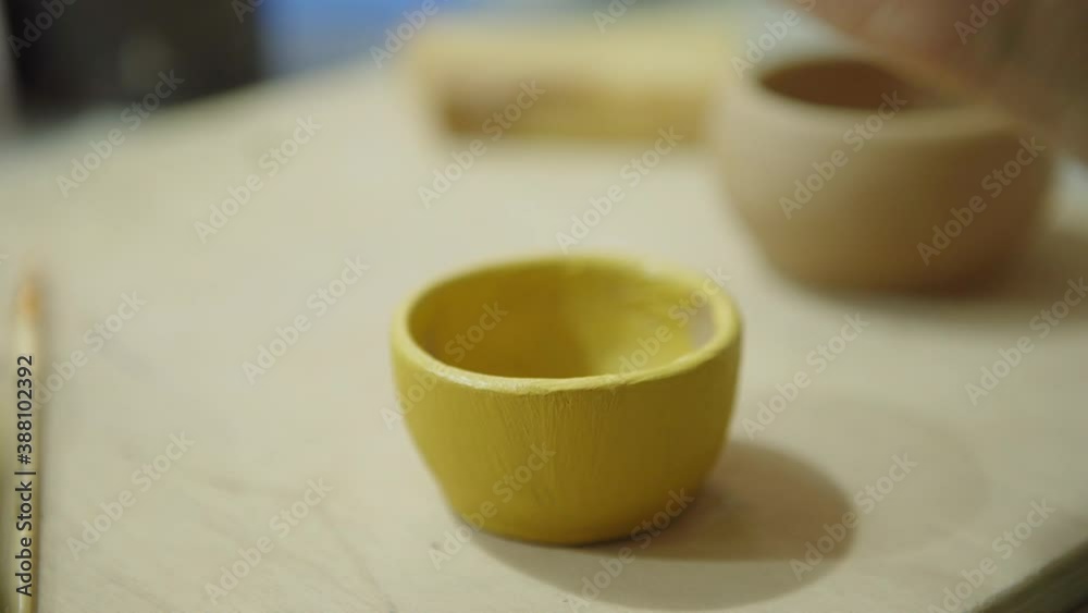 Closeup of hand of female artist using paintbrush to color freshly made small ceramic bowl in yellow Closeup of hand of female artist using paintbrush to color freshly made small ceramic bowl in yellow