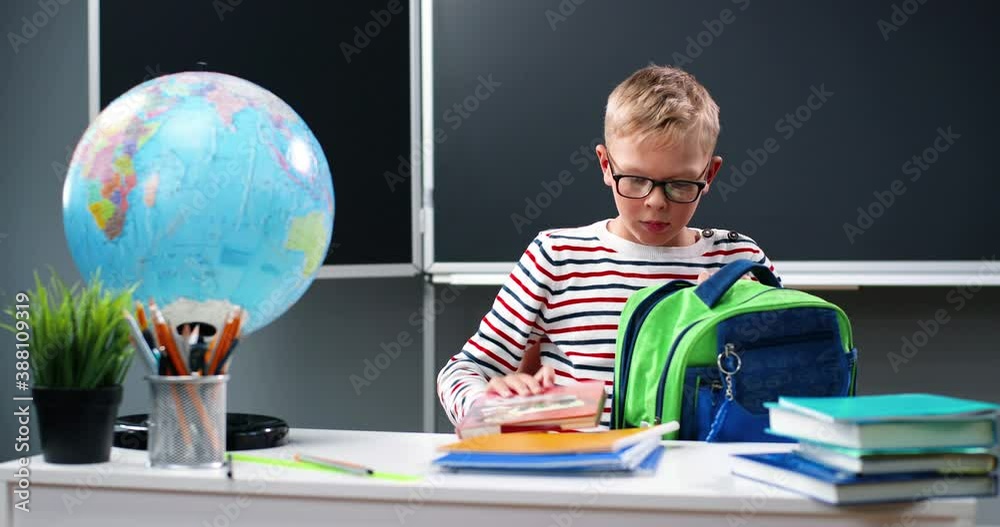 Caucasian teen boy in glasses putting things in backpack from desk in room and preparing to school. Schoolboy in eyeglasses getting ready to classes at home. Schooling life. Caucasian teen boy in glasses putting things in backpack from desk in room and preparing to school. Schoolboy in eyeglasses getting ready to classes at home. Schooling life.