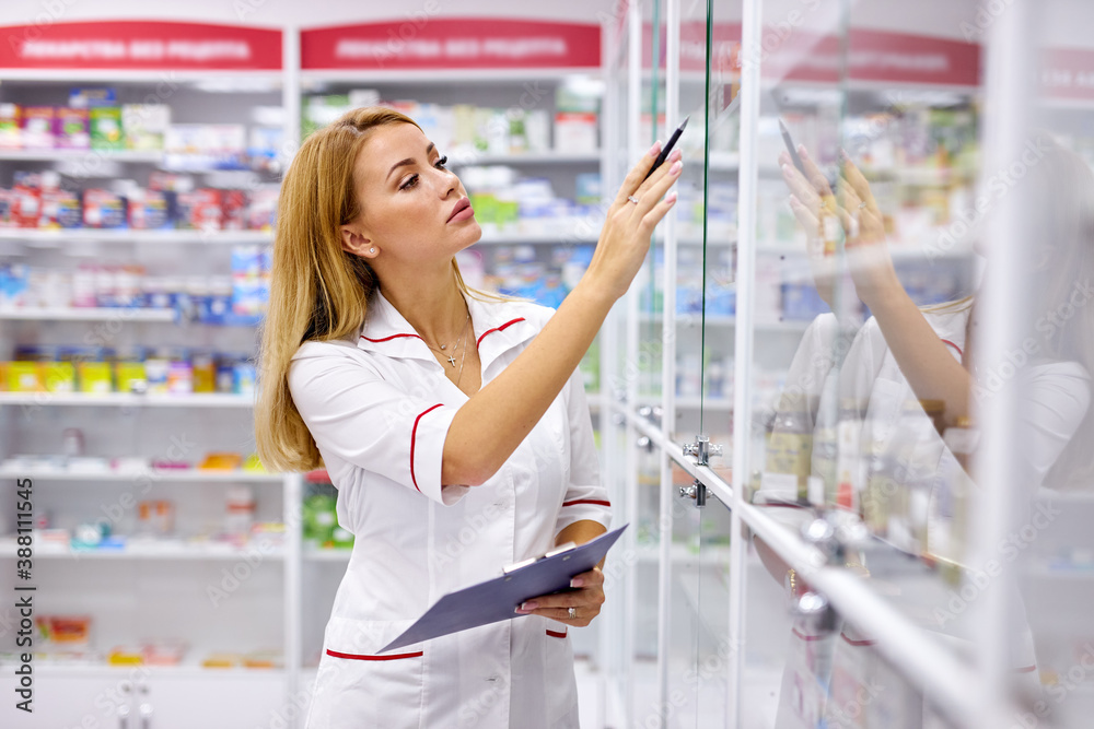 portrait of good-looking blonde pleasant female pharmacist writing on clipboard at pharmacy, make notes, wearing white medical gown. check list of drugs