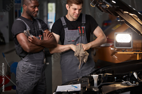 multi-ethnic team of african and caucasian men working in auto service together, friendly men in uniform repair the car hood