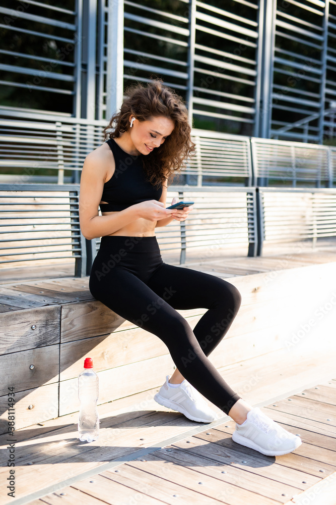Sunny summer day. Two young women athletes in sports clothes are sitting on bench, relax after sports training, use smartphone checking email, browsing internet, chatting.