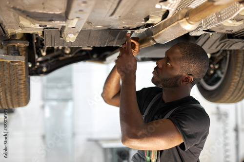 фотографія young male car mechanic in uniform checking car in automobile service with lifted vehicle, handsome hardworking guy working under car condition on lifter
