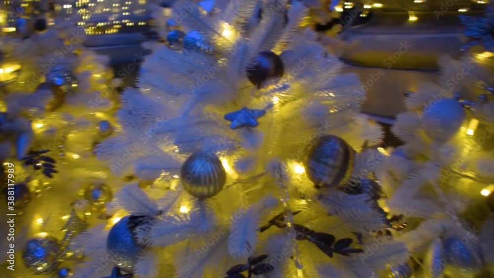 White Christmas tree  with silver decorations and golden bulbs ball decoration hanging in the night city.  Camera, defocused. Celebration concept, festive atmosphere. Night Street Of The City