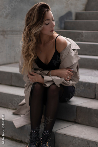 attractive slim young brunette in fashionable trench and black dress sitting on grey concrete staircase. urban lifestyle concept. © OliaVesna
