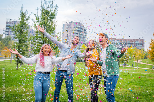 Group of stylish friends smiling and enjoying party throwing colorful confetti to the sky - Multietnic guys in the park throwing colorful confetti in the air