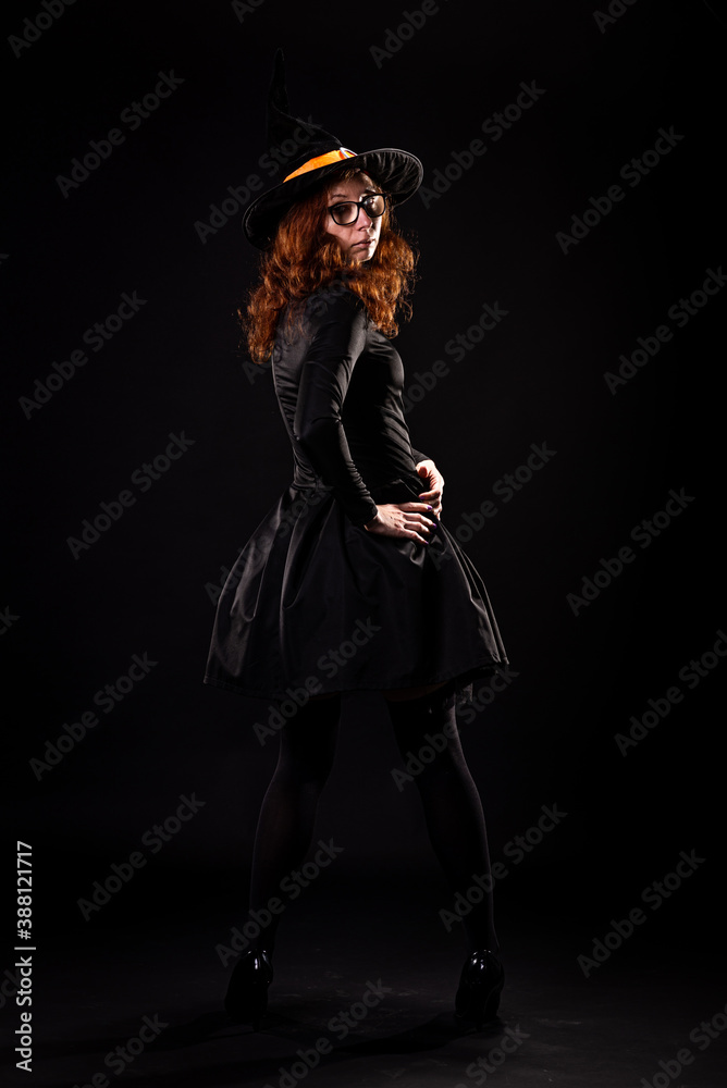 Young woman in black dress and witch hat on a black background, back view. Young woman in black dress and witch hat on a black background, back view.