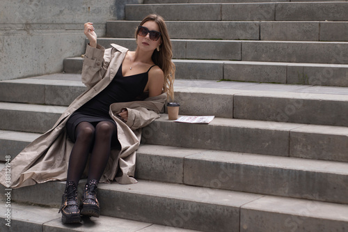 attractive slim young brunette in fashionable trench and black dress sitting on grey concrete staircase near a brown paper cup and a newspaper. © OliaVesna