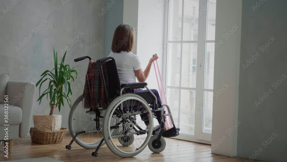 Rear view of young disabled woman in wheelchair exercising with resistance band. Training, sport, recovery and lifting, stretching exercise at home