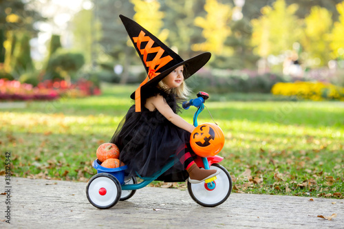 cute little girl in a witch's costume rides down the street on a children's bike with pumpkins and candy © КРИСТИНА Игумнова