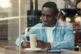 Concentrated black male entrepreneur uses smartphone while sitting in cafe with coffee near window.