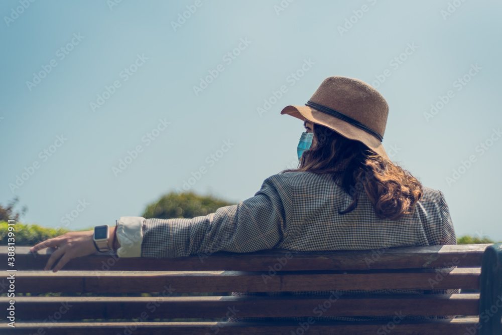 Retrato de una mujer con sombrero sentada de espalda en un banco al aire  libre con una mascarilla medica. foto de Stock | Adobe Stock