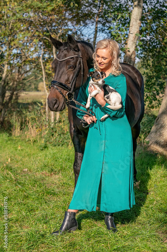 Beautiful blond smiling girl with her horse and dog in the forest. Wearing a green dress. Selective focus