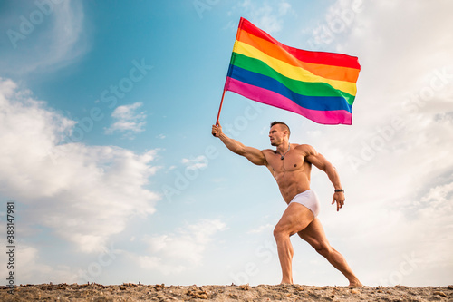 Shirtless young man holding rainbow flag walking at beach against sky photo