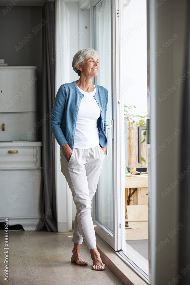 Senior woman standing with hands in pockets while looking out of window at home Senior woman standing with hands in pockets while looking out of window at home