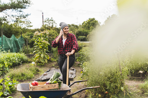Fotografie Young woman talking over mobile phone while standing by wheelbarrow in vegetable