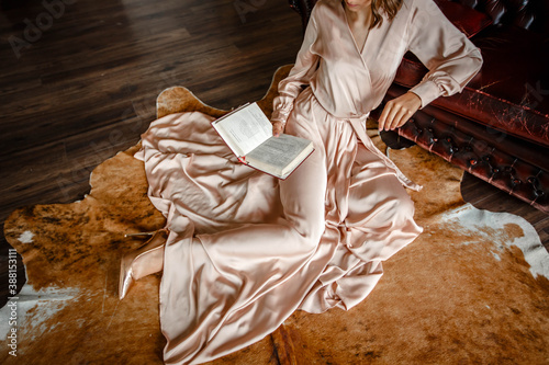 Girl in a long dress sits on the floor by a leather sofa and reads a book. Luxury interior. cropped photo