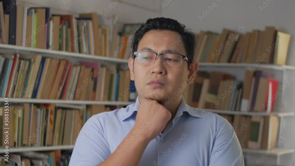 Asian man thinking while standing in front of bookshelf, education concept, studying in library, serious expression Asian man thinking while standing in front of bookshelf, education concept, studying in library, serious expression