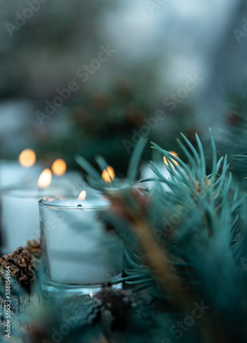 five lit white votive candles burning flames amid green pine branches and decorative pine cones