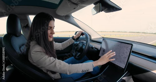 Beautiful Caucasian young woman sitting in electric vehicle and typing on big touch screen. Cheerful pretty female driver tapping on-board computer in car and searching route on GPS navigation map photo