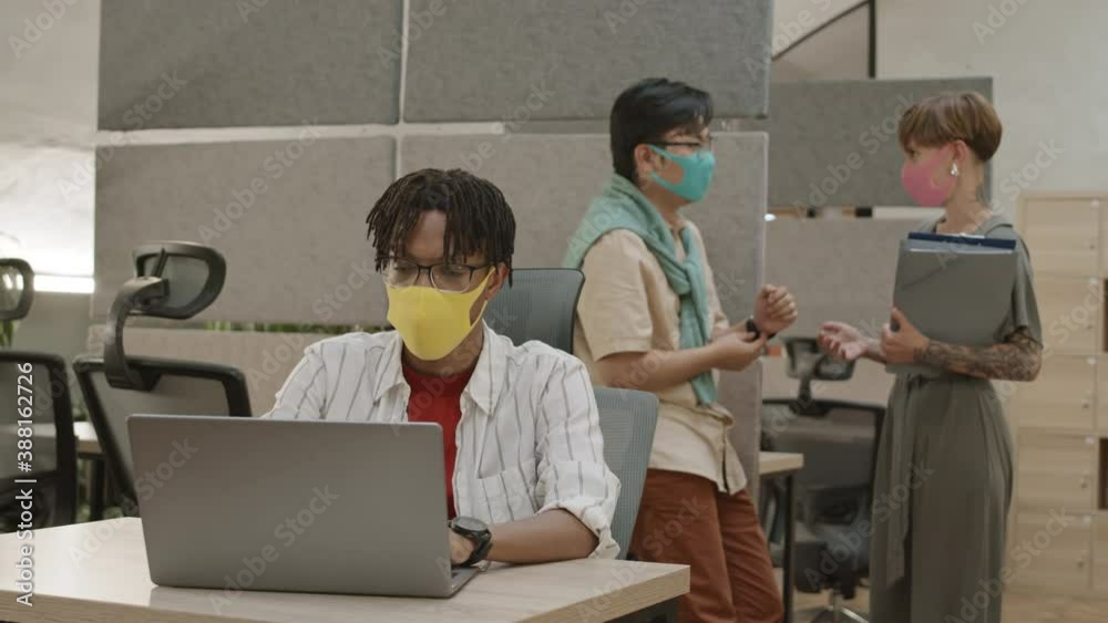 Medium shot of young African-American man wearing mask sitting at workplace, working on computer while his two diverse coworkers wearing masks finishing talk and then going away Medium shot of young African-American man wearing mask sitting at workplace, working on computer while his two diverse coworkers wearing masks finishing talk and then going away