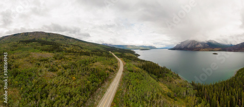 Beautiful Panoramic View of Road alongside Scenic Lake surrounded by Mountains and Trees on a Cloudy Day. Aerial Drone Shot. Taken near Klondike Highway  Southern Yukon  Canada.