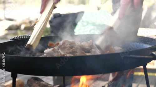 Close up shot of man stir-frying pieces of beef kebab meat in a muurikka griddle pan, over a hot wood fire in a camping environment   photo