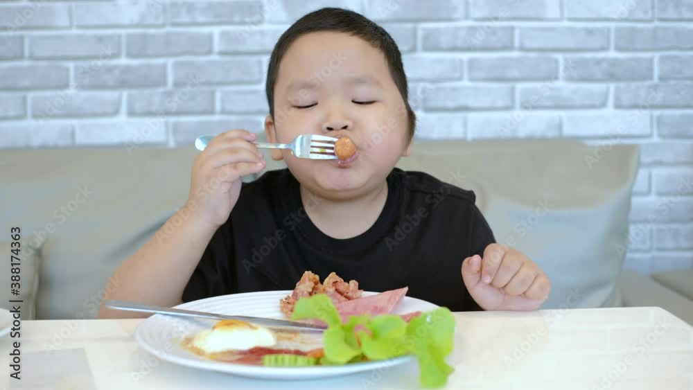 Little Asian boy eating morning breakfast with fried egg, bacon, potatoes and sausage at home. Little Asian boy eating morning breakfast with fried egg, bacon, potatoes and sausage at home.