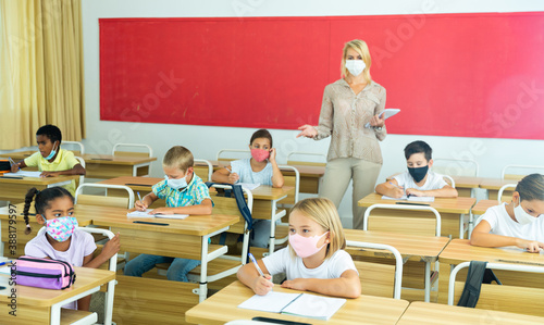 Multiracial group of kids wearing face masks working at class, writing and listening explanations of teacher in classroom