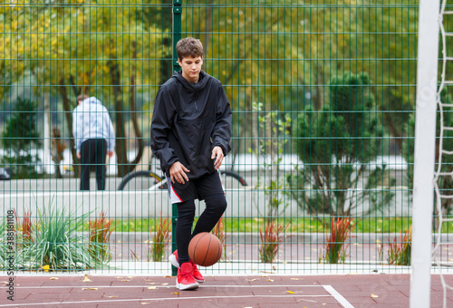 Cute young boy plays basketball on street playground. Teenager with orange basketball ball outside. Hobby, active lifestyle, sport activity for kids.