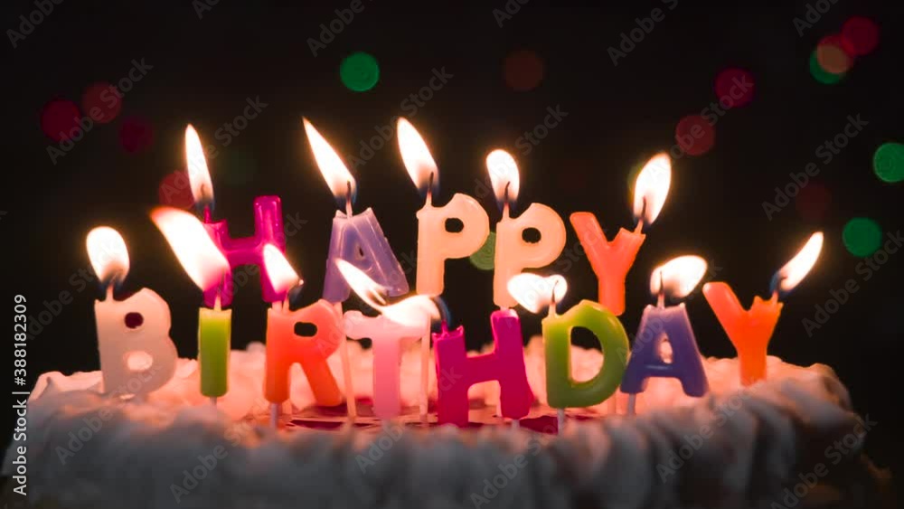 Fluttering Holiday Candles on the Cake. Cake with candles in the form of an inscription Happy Birthday. In the background, flashing and moving lights bokeh in the dark Fluttering Holiday Candles on the Cake. Cake with candles in the form of an inscription Happy Birthday. In the background, flashing and moving lights bokeh in the dark