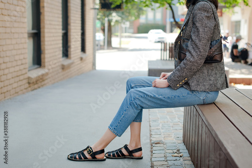 Young fashionable woman wearing gray tweed jacket with golden buttons, blue jeans, sweater, black satin fisherman sandals and holding black leather baguette handbag. Street style.