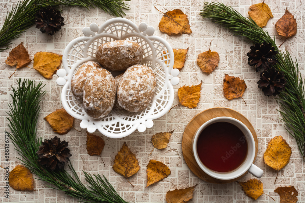 Black tea cup on wooden saucer with white wicker basket of gingerbreads in natural frame of pine branches and pine cones against pastel shades background with fall leaves Black tea cup on wooden saucer with white wicker basket of gingerbreads in natural frame of pine branches and pine cones against pastel shades background with fall leaves
