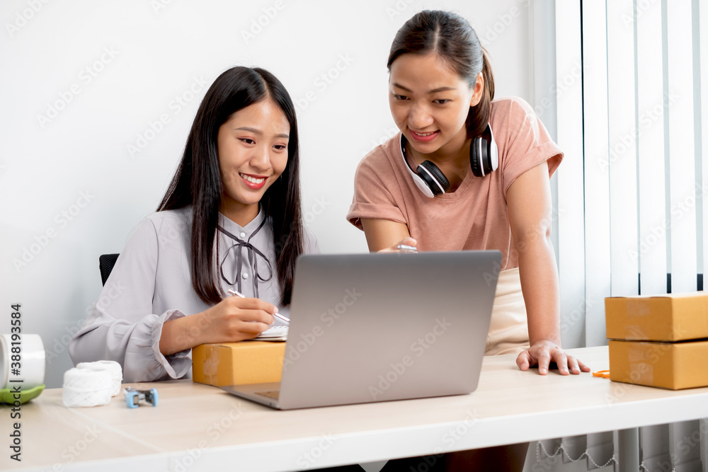 Asian woman business two people owner with many parcel boxes on the table happy online sales job, use your laptop, get an order from customers, take notes, and make arrangements for delivery by post Asian woman business two people owner with many parcel boxes on the table happy online sales job, use your laptop, get an order from customers, take notes, and make arrangements for delivery by post