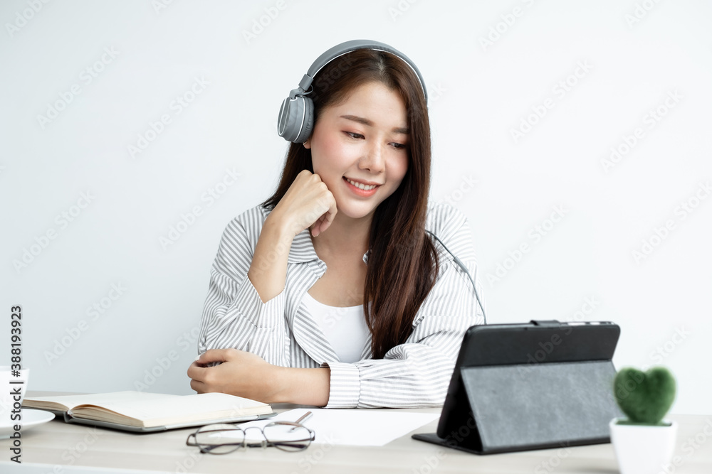 Video Calling a young Asian woman wearing headphones communicates via online long-distance video on a digital tablet happily greetings with a friend Video Calling a young Asian woman wearing headphones communicates via online long-distance video on a digital tablet happily greetings with a friend