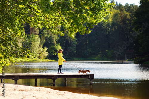 Portrait of a girl in a yellow jacket on a wooden bridge near the lake with a pet red Dachshund on a leash