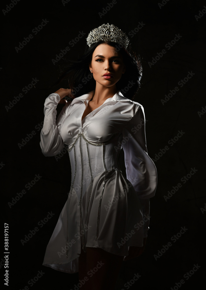 Gorgeous rich woman princess in crown and white corset mini dress has turned to look at camera over dark background