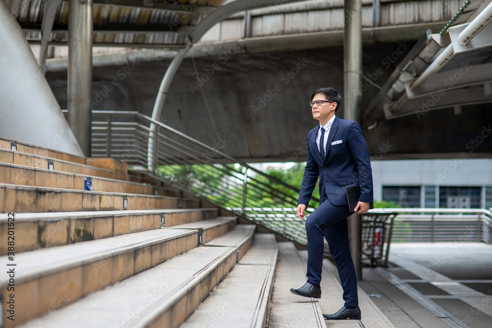 Confidence Asian businessman office worker in suit holding digital tablet and walking up the stairs at railway station in the city. Transportation, technology and financial business concept.