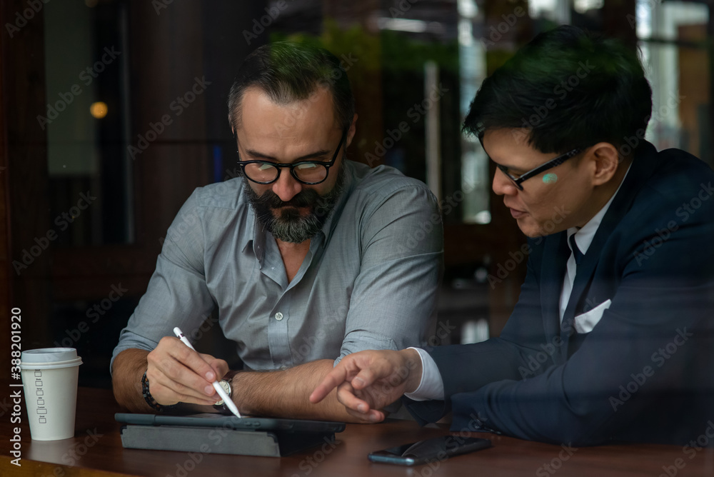 Caucasian beard man creative freelance sitting in coffee shop with Asian businessman in suit discuss about business plan with using digital tablet. Business meeting teamwork and technology concept. Caucasian beard man creative freelance sitting in coffee shop with Asian businessman in suit discuss about business plan with using digital tablet. Business meeting teamwork and technology concept.