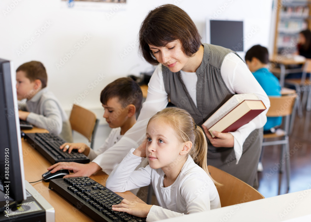Fototapeta premium Friendly woman teacher helping tween girl during lesson in computer room of school library ..