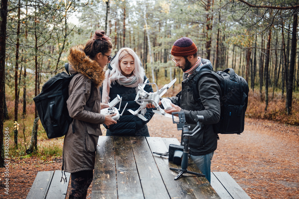 Company of three persons a bearded guy and two beautiful girls they tuning and preparing quadcopter at table in autumn forest.