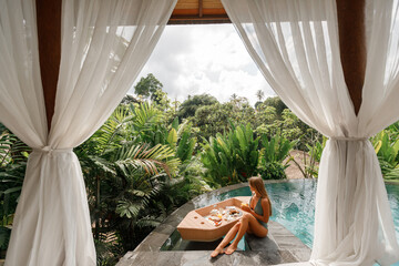 Woman enjoying breakfast in outdoor daybed with side curtains next to the swimming pool, the perfect begining of summer day