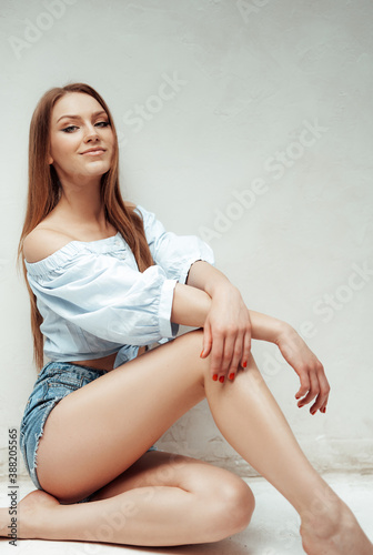 Attractive and young woman with brown hairs in white shirt poses near wall on floor in white room.