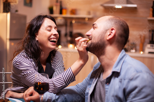 Husband feeding wife with grapes during romantic dinner celebrating relationship. Wife and husband celebrating anniversary with red wine, tender moments at candle lights in kitchen.