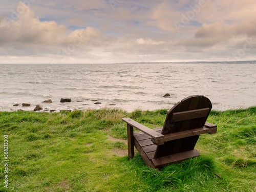 One empty wooden chair on a grass by ocean with magnificent view. Cloudy sky over calm water surface. Concept enjoying nature and relaxation.