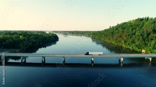 Tracking aerial shot of semi truck driving on bridge over river during sunrise with light streaks and fog on the water followed by other trucks.