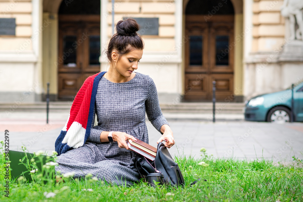 Obraz premium Young adult female student puts books to backpack sitting on grass near university building