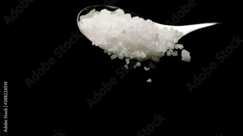 Full spoon of white sea salt. Coarse rock salt mineral grains falling from spoon in slow motion isolated on black background. Food, healthy eating, halite, nutrition, harm benefits of salt concept